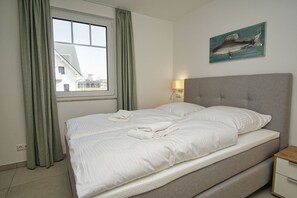 Una cama bien hecha con ropa de cama y toallas blancas, un cabecero gris, una lámpara de noche y una obra de arte en la pared con un pez.