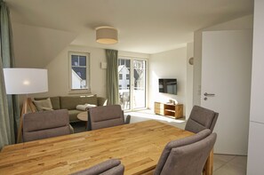 A modern living room with a wooden dining table, grey armchairs, a sofa, a TV, and a window with curtains. - Beachfront vacation rental with a balcony, pool access, and a sauna - Rex Rugia - Rex Rugia (Göhren)