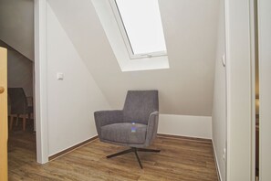 Une petite pièce moderne avec un plafond en pente et un puits de lumière, comprenant un fauteuil recouvert d’un tissu gris et un plancher en bois.