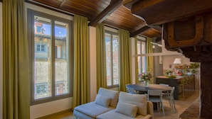 A cozy living room with a sofa, dining table, and chairs. Large windows with curtains provide natural light and a view of the outside.