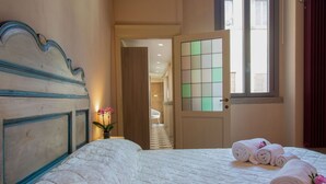 Una habitación de hotel con cama, puerta con panel de vidrio esmerilado, ventana con cortinas y pasillo que conduce a otra habitación.