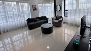 A modern living room with a black sofa, a grey armchair, and a coffee table. There is a flat-screen TV on a stand and a large window with sheer curtains. - Comfy Arte Plus Ampang City 7PAX-3BR-2CP (Kuala Lumpur)