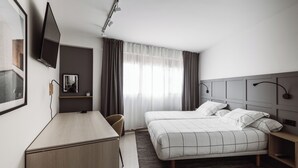 Une chambre d’hôtel comprenant un lit, un bureau, une chaise, un téléviseur fixé au mur et une fenêtre avec des rideaux.