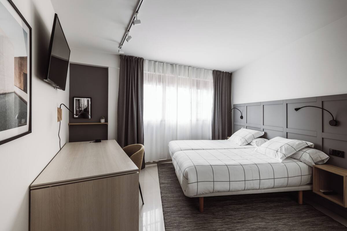 A hotel room with a bed, a desk, a chair, a TV mounted on the wall, and a window with curtains.