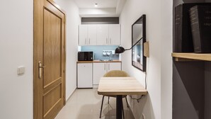 Un couloir étroit mène à une cuisine dotée de meubles blancs, d’un four à micro-ondes et d’un évier. On y trouve une table avec une chaise et une lampe noire. Des livres sont rangés sur une étagère, à droite.