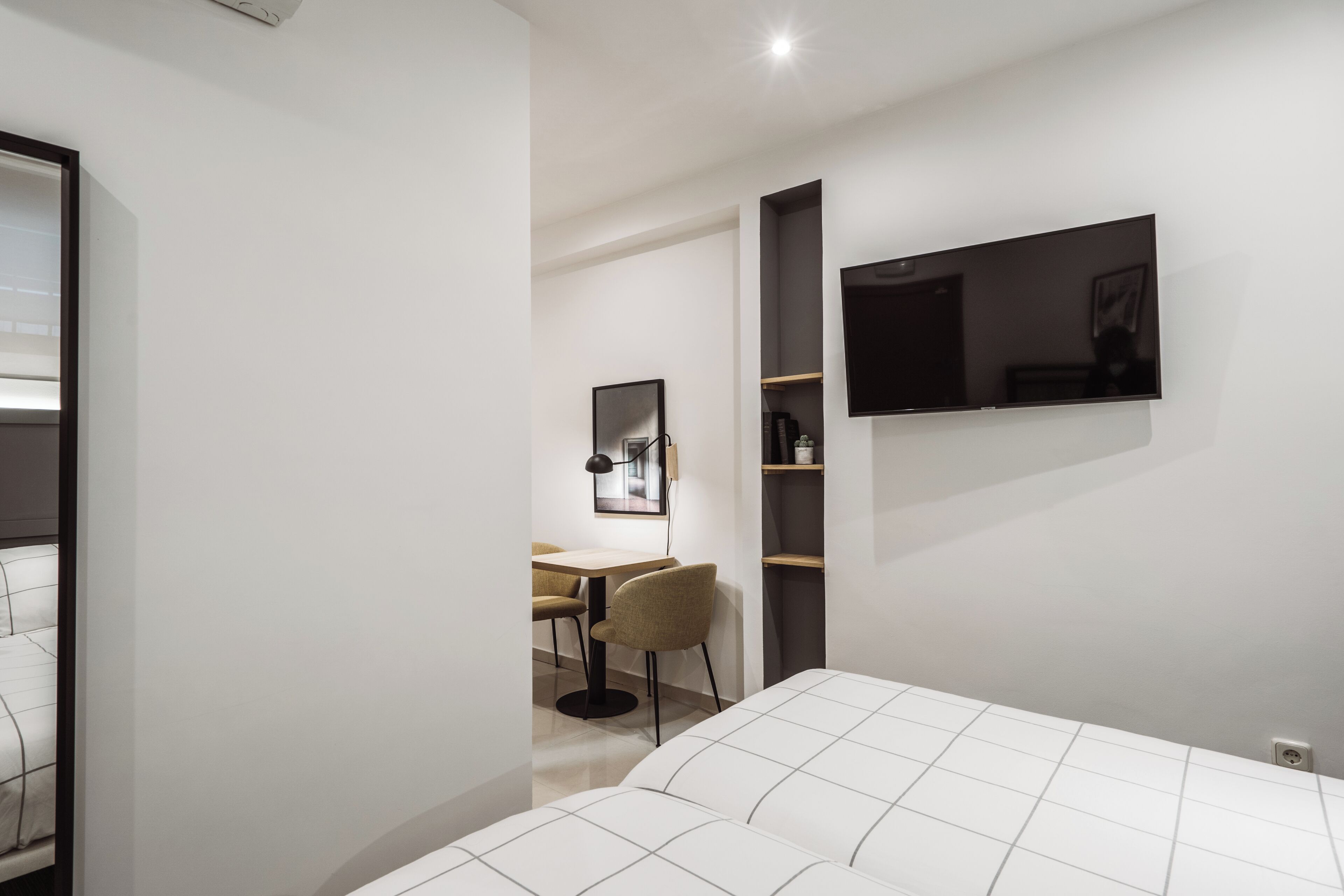 A modern hotel room with a large bed, a desk with a chair, a TV mounted on the wall, and a small shelf with items.