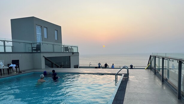 Una piscina con vistas a la puesta de sol sobre el océano, barandilla de cristal y estructura de edificio moderna.