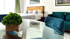 Habitación de hotel con una cama, un sofá verde, una mesa de vidrio con una planta y dos cuadros en la pared.