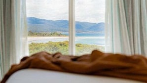 Seng med udsigt til strand og bjerge gennem et vindue med gardiner.