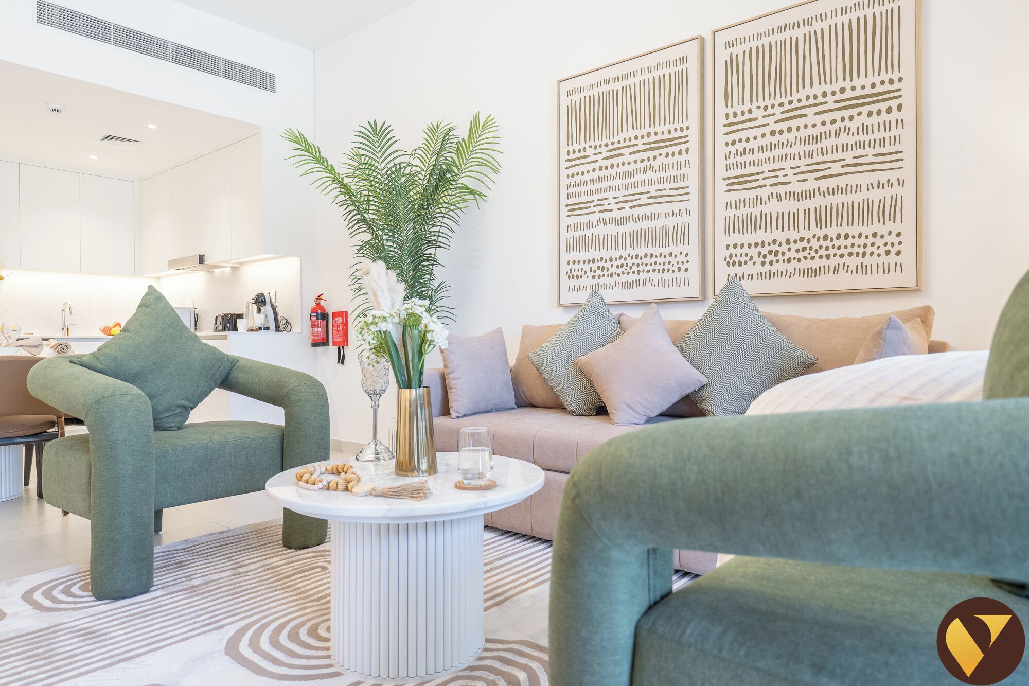 Un salon moderne avec des fauteuils verts, un canapé beige et une table basse blanche agrémentée d’objets décoratifs. On y trouve également des œuvres d’art murales abstraites et une plante en pot.