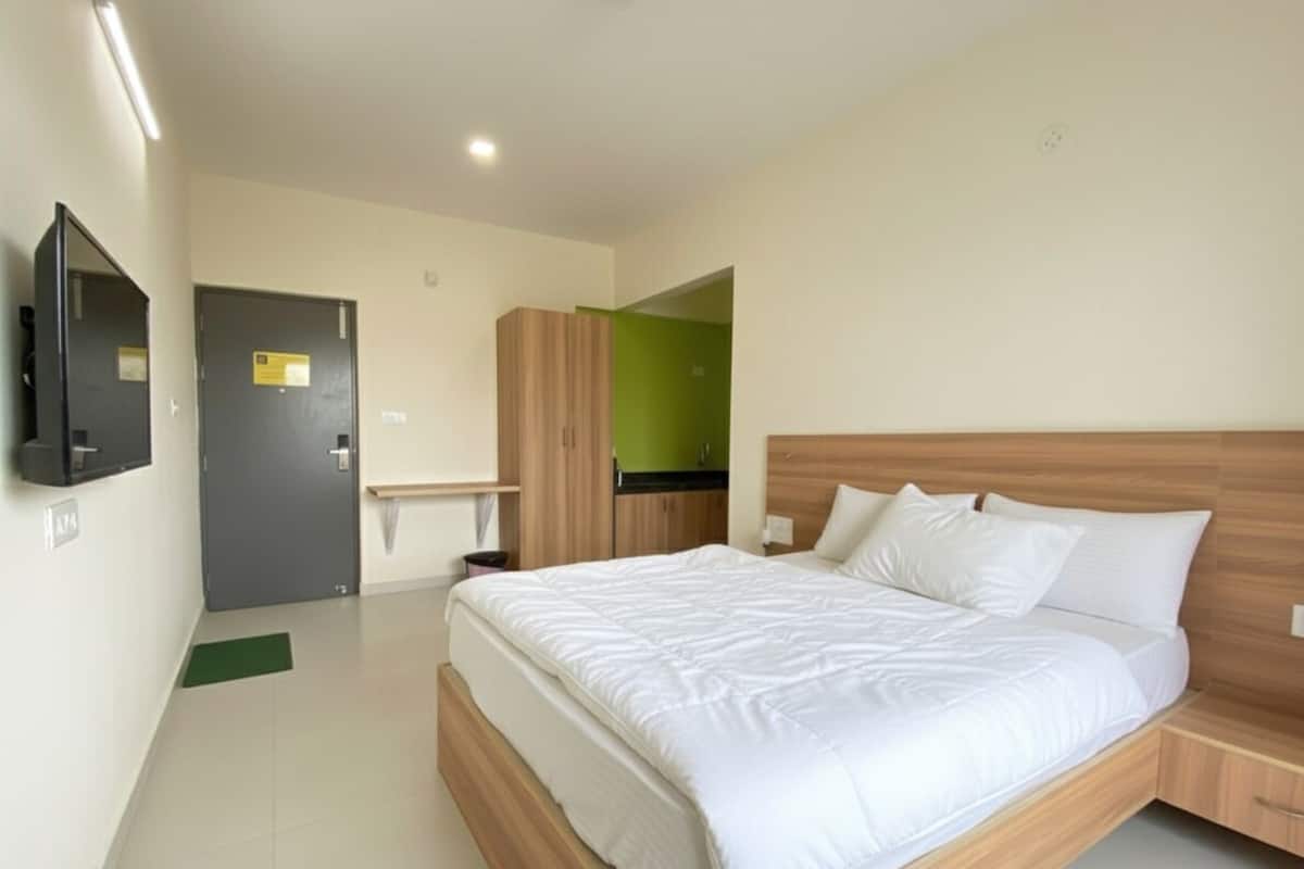 A modern hotel room with a large bed, a wooden headboard, a flat-screen TV mounted on the wall, a green mat on the floor, and a door with a sign.