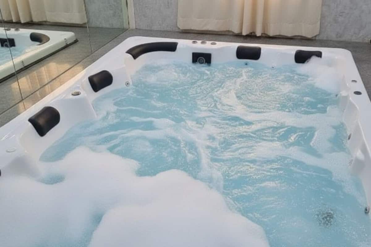 A hot tub with a rectangular shape and multiple jets, situated indoors with a tiled floor and a window with curtains.