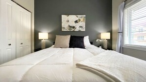 Een net opgemaakt bed met witte lakens, twee bedlampjes, een abstract schilderij aan de muur en een raam met gordijnen.