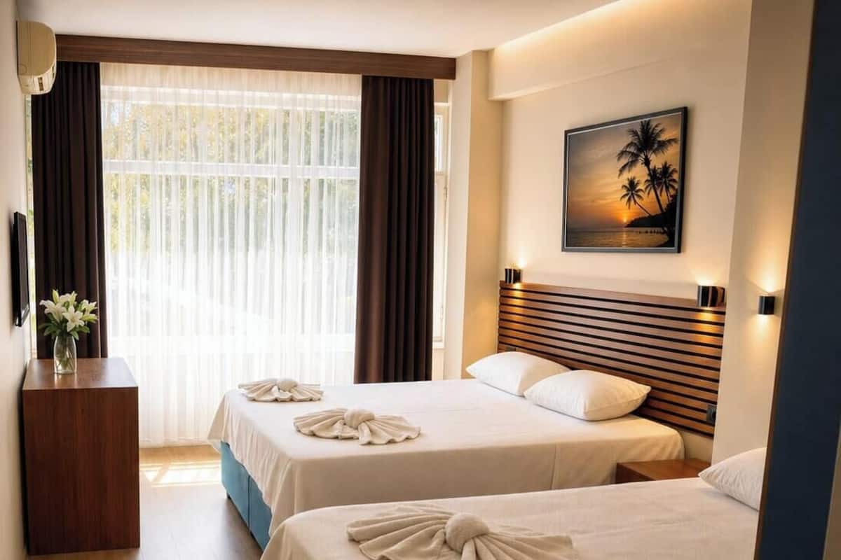A hotel room with two beds, a wooden headboard, a painting of palm trees, and a desk with a vase of flowers.