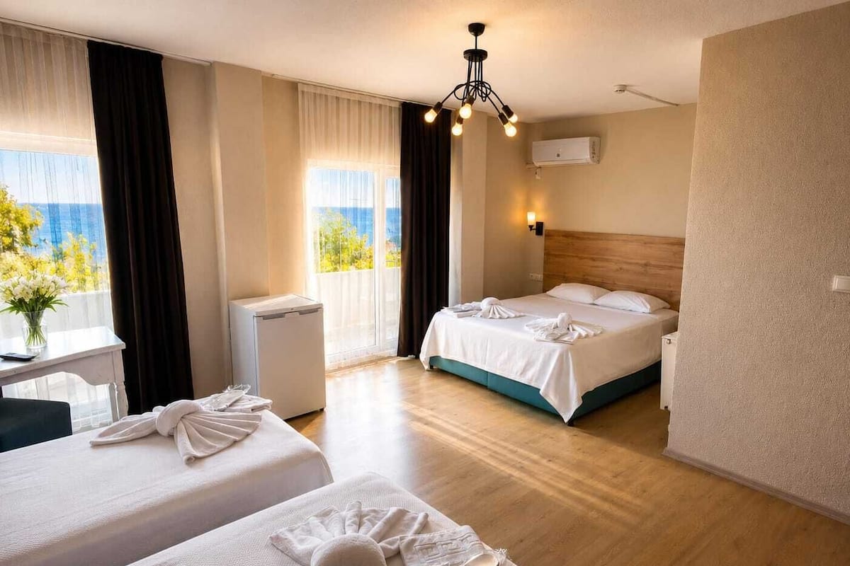 A hotel room with two beds, a desk, a chair, a window with curtains, an air conditioning unit, and a view of the sea.