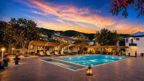 A pool area with a clear blue pool, surrounded by buildings with a thatched roof and a white building with a blue roof. - Blue Park Assos (Ayvacik)
