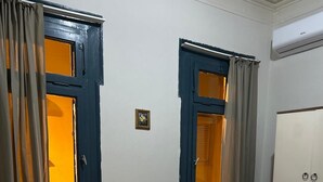 A single bed with a grey mattress and white sheets, a small bedside table with a lamp, a wardrobe with two doors, and two windows with curtains. - Arada Ayvalık (Ayvalik)