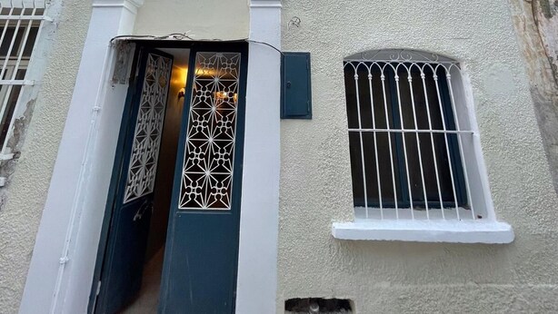 A two-story building with a white facade, a central entrance with a decorative metal gate, and barred windows. - Arada Ayvalık (Ayvalik)