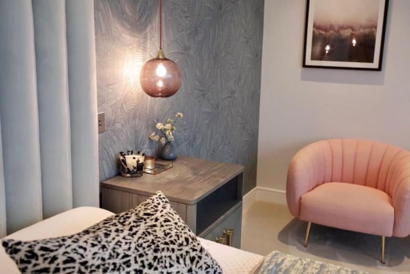 Een slaapkamer met een bed, nachtkastje, een roze stoel, een hanglamp en een lijst met een foto aan de muur.