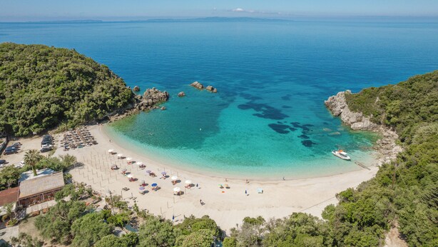 Vue aérienne d’une plage aux eaux turquoise et limpides, avec des formations rocheuses et un bateau amarré près du rivage.