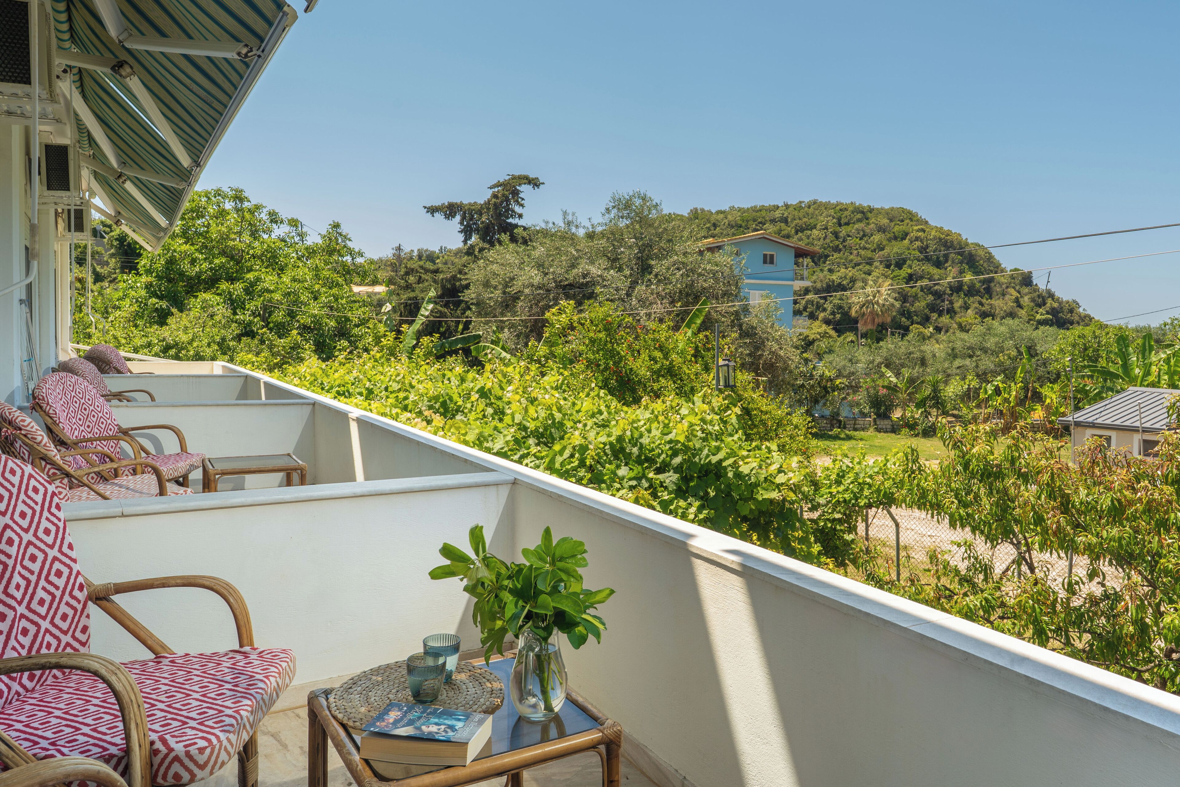 A balcony with wicker furniture, a table with a vase and books, and a view of a hillside with houses and greenery.