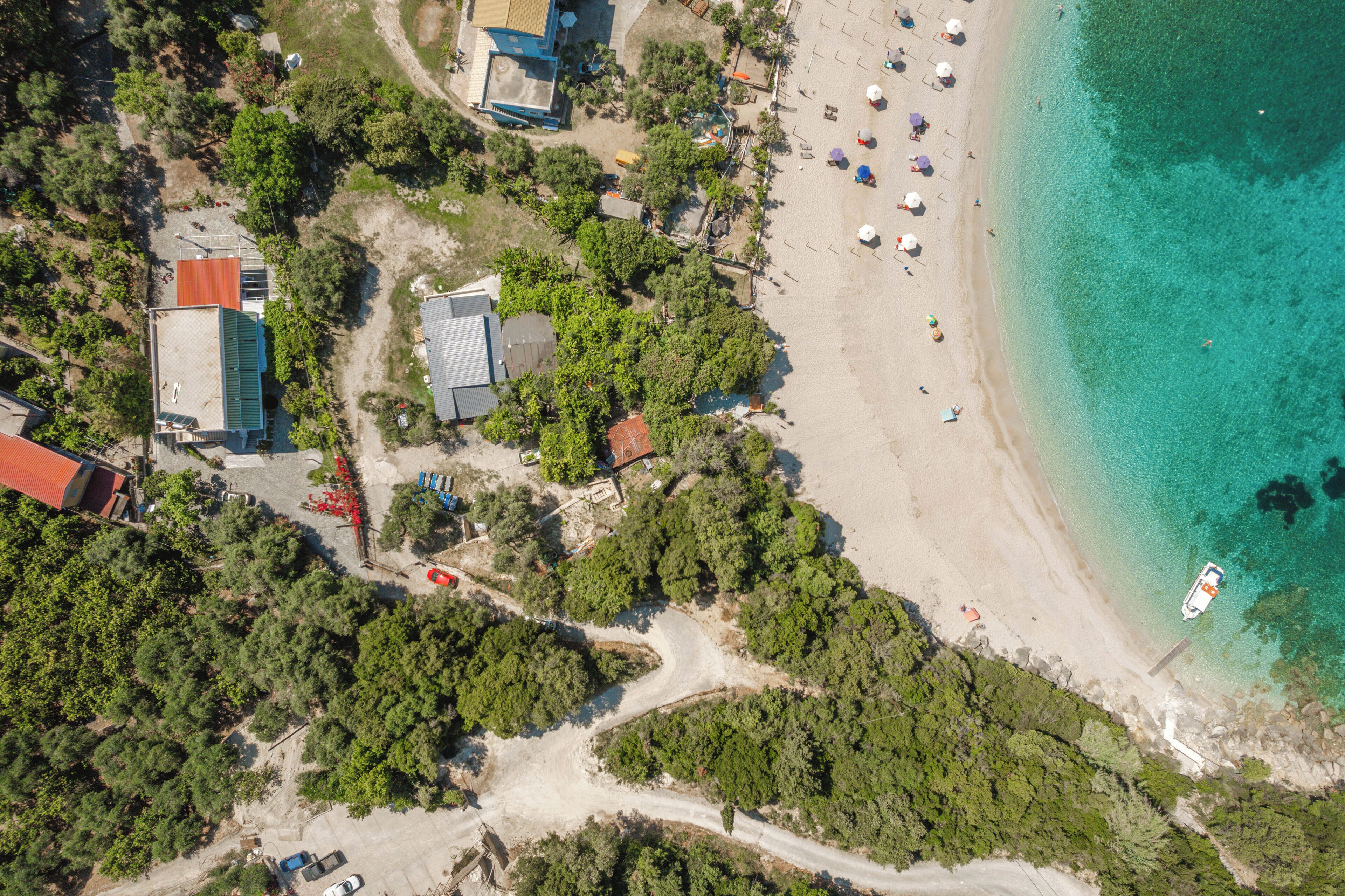 An aerial view of a beach with clear turquoise waters, a sandy shore, and a few buildings surrounded by dense greenery.