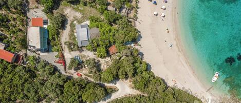 An aerial view of a beach with clear turquoise waters, a sandy shore, and a few buildings surrounded by dense greenery.