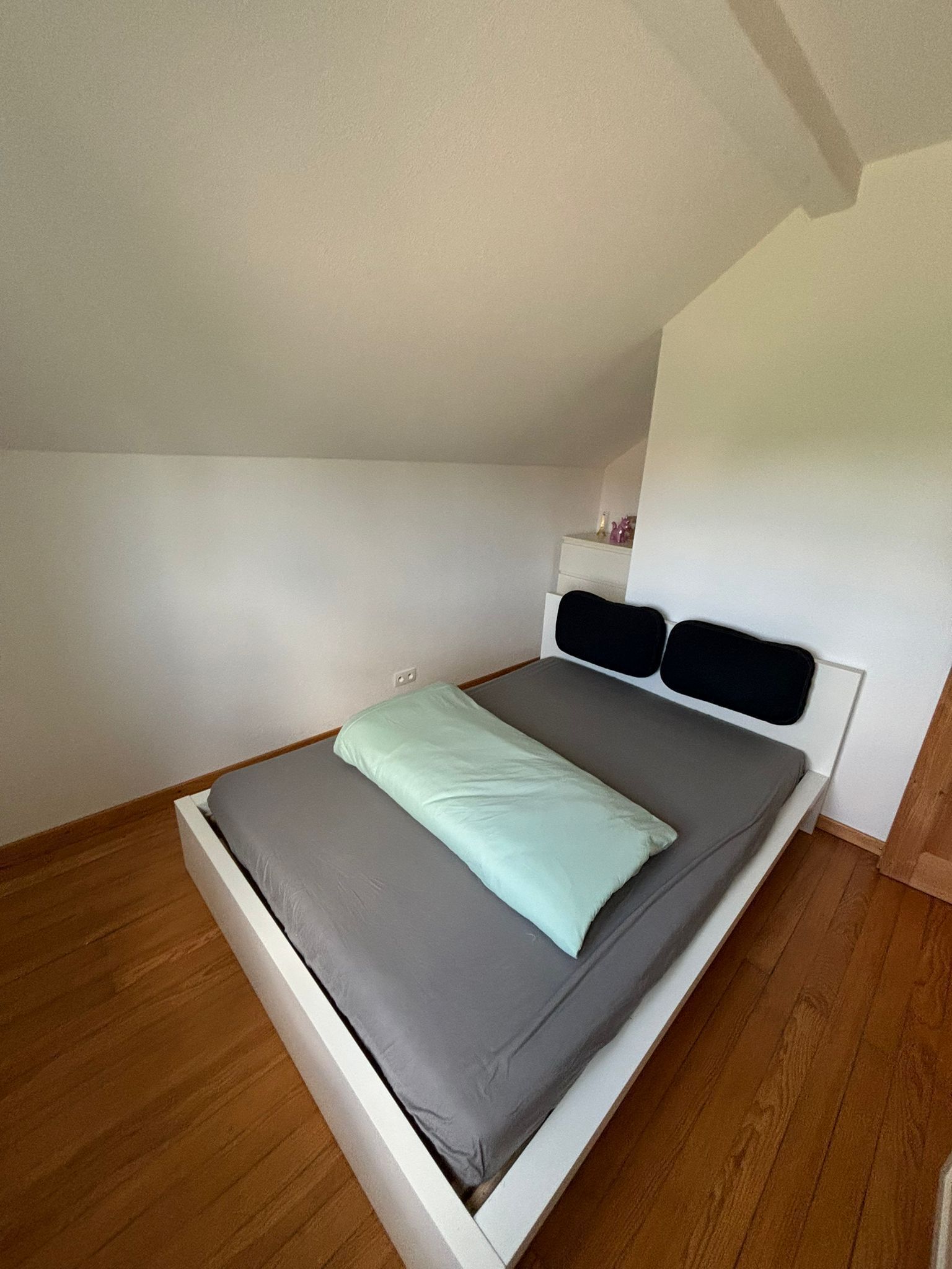 Ein Einzelbett mit grauem Lattenrost und einer hellgrünen Decke, ein weißes Kopfteil und ein Holzboden.