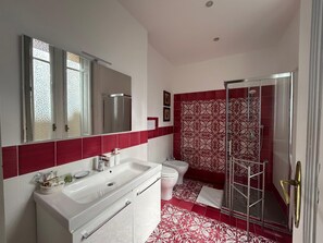 A modern bathroom with a white vanity, a glass shower enclosure, and a red-tiled shower area. - La Giulietta- Penthouse (Salerno)