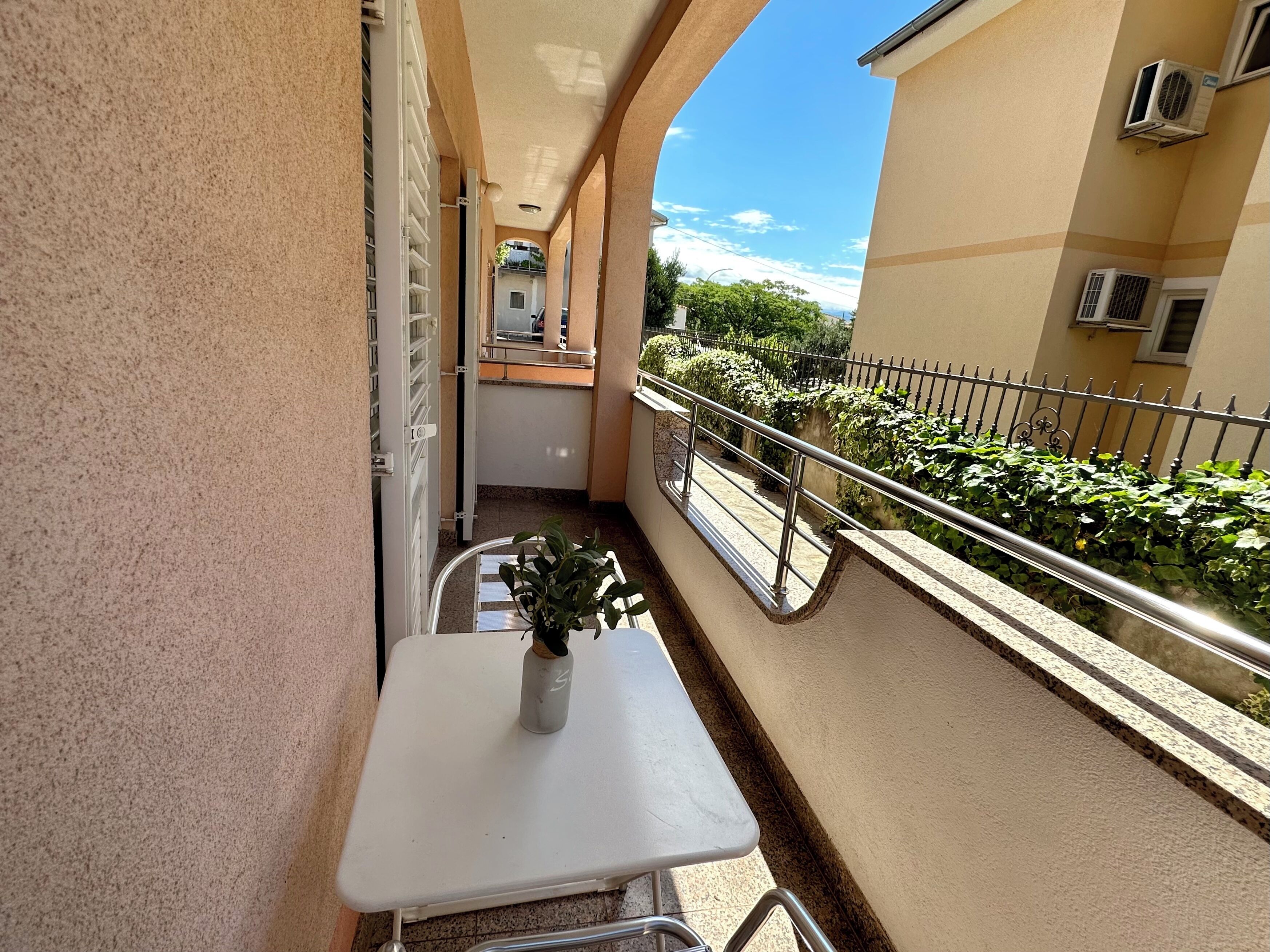 A balcony with a small table and a potted plant, a railing, and a view of a building and greenery.
