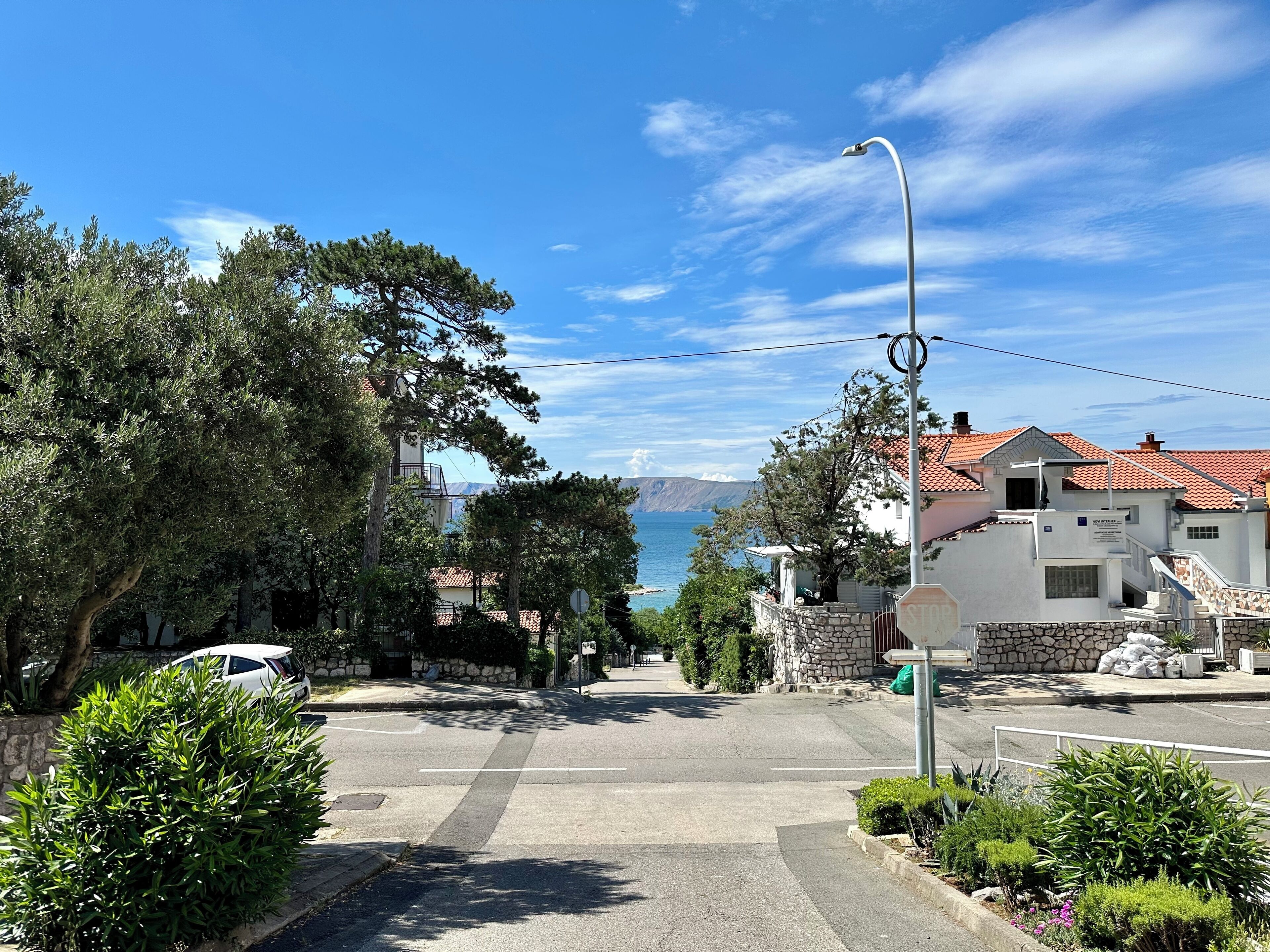 A coastal street with a crosswalk, a stop sign, and a view of the sea and mountains in the distance.