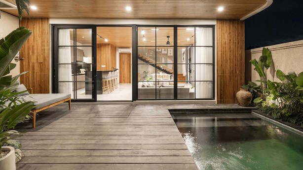 Ein modernes Haus mit einer Holzdachterrasse, einem Pool und einer Glastür, die zu einem Innenbereich mit Treppe und Küchenbereich führt.