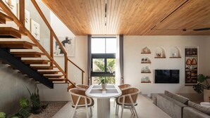 Ein modernes Interieur mit einer Holztreppe, einem Essbereich mit Korbstühlen, einem grauen Sofa und einem großen Fenster, das natürliches Licht hereinlässt.