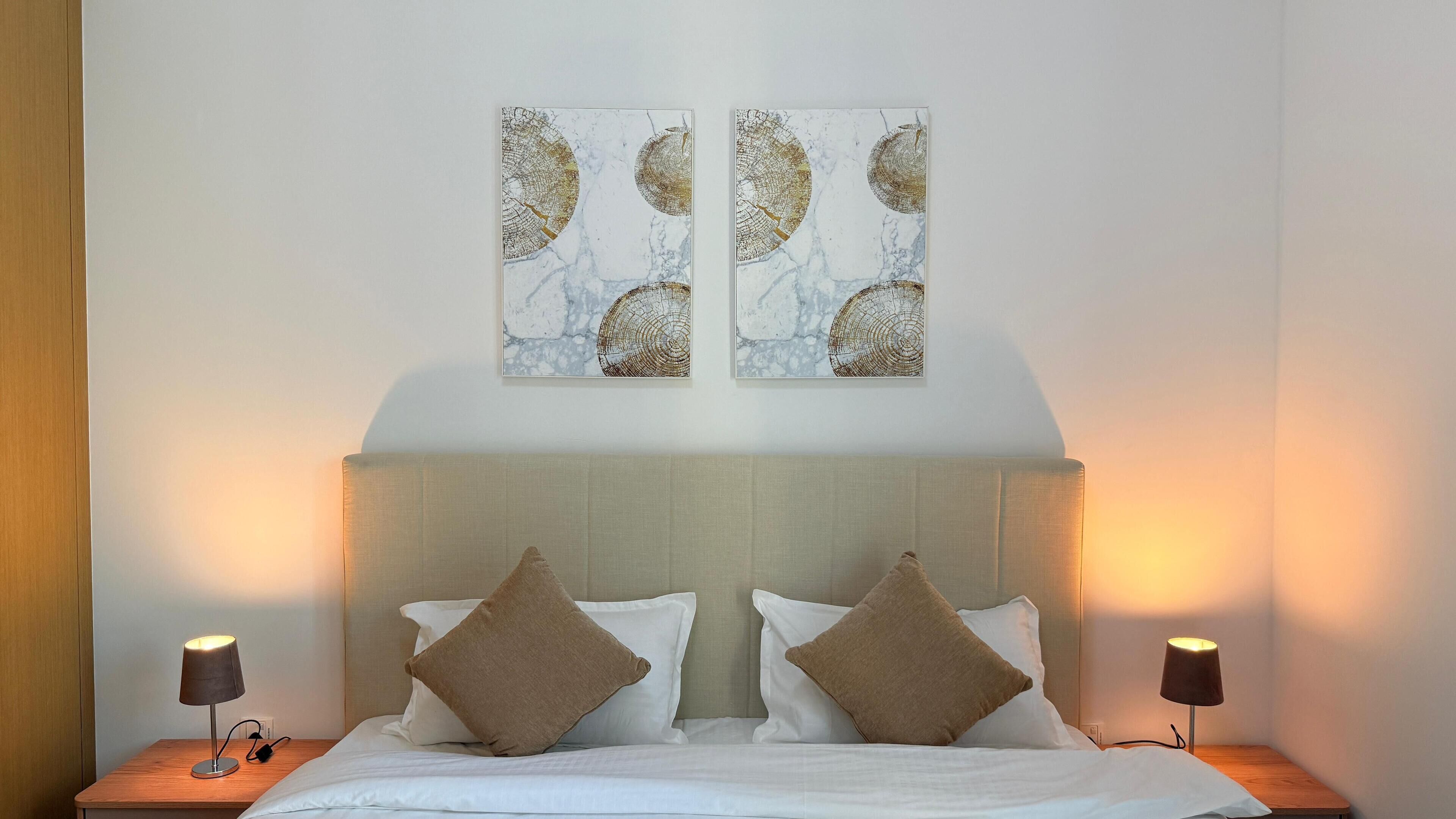 Quarto de hotel com cama, duas mesinhas de cabeceira, duas luminárias e duas obras de arte penduradas na parede.