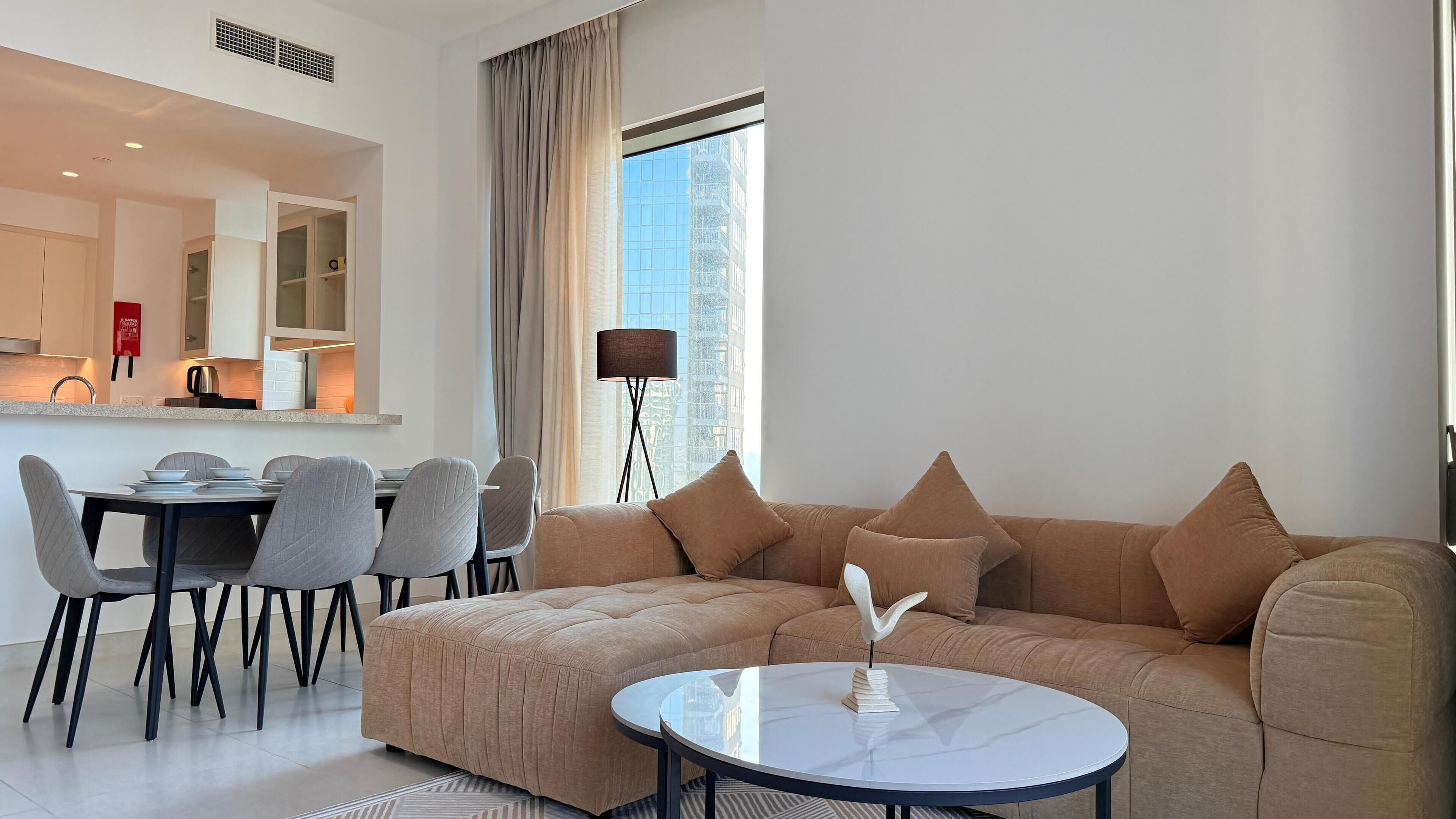 A modern living room with a sectional sofa, a dining table with chairs, a round coffee table, and a large window with a view of buildings.