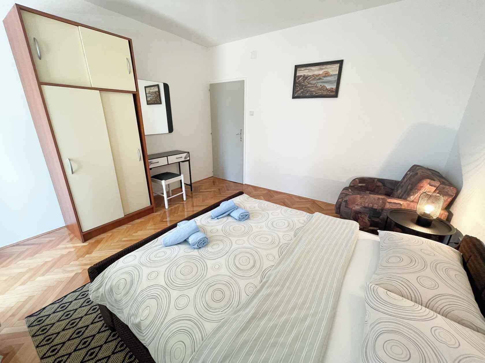 A bedroom with a bed, a desk, a chair, a wardrobe, and a small table with a lamp.