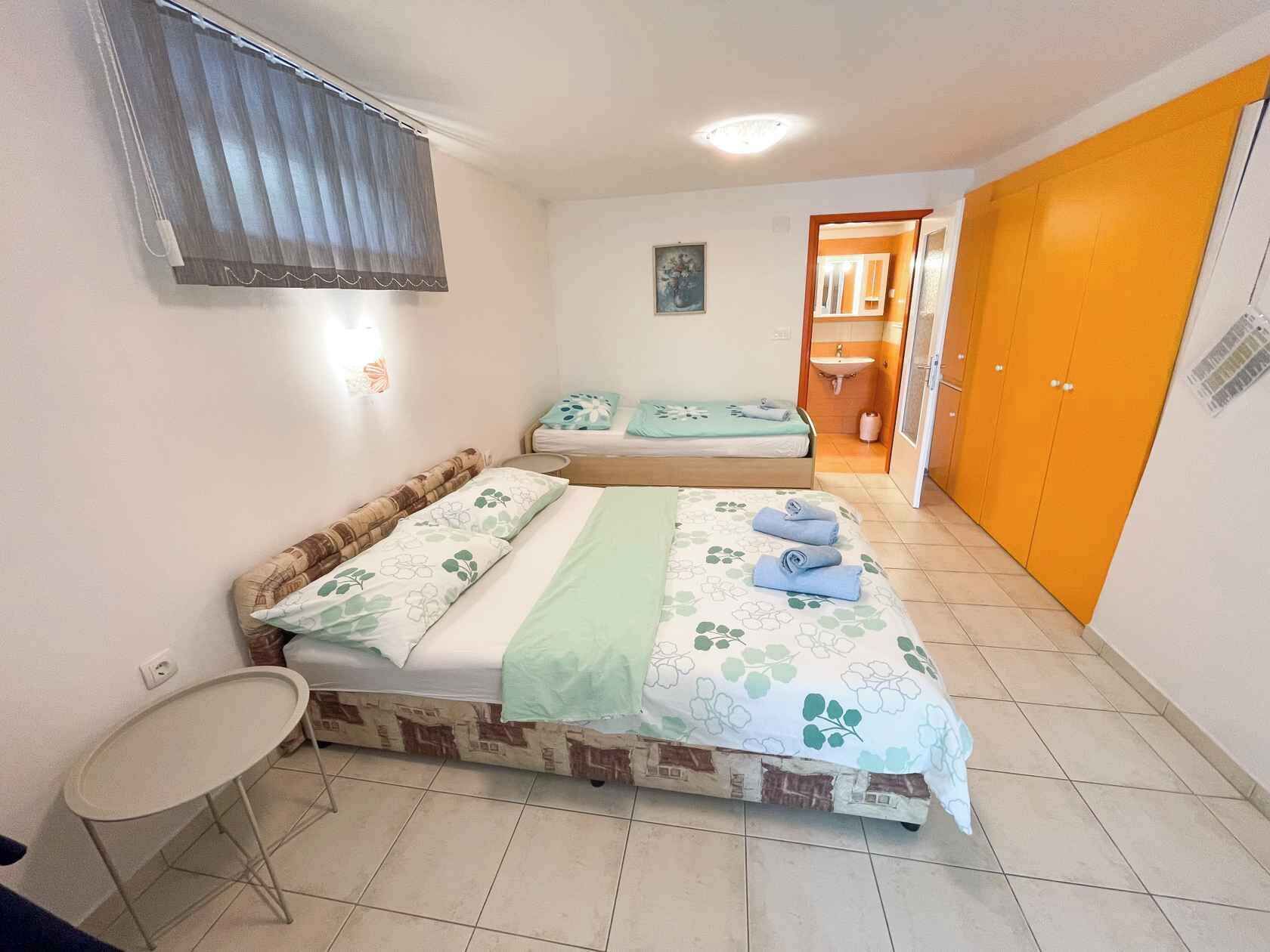 A room with two beds, a small table, a window with blinds, a bathroom with a sink, and an orange door.