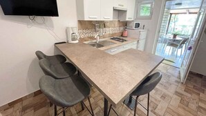 A compact kitchen with a flat-screen TV, a coffee maker, a microwave, and a gas stove. There are three bar stools and a small dining area with a table and chairs. - Apartmani Talia Crikvenica (Crikvenica)