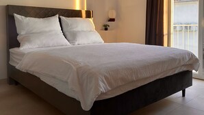 Ein ordentlich bezogenes Bett mit weißen Leinen in einem Zimmer mit einem großen Fenster, einem Nachttisch mit Lampe und Blick auf ein Gebäude.