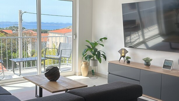 Ein modernes Wohnzimmer mit einer Couch, einem Couchtisch und einem Fernseher auf einem Holzschrank. Ein Balkon bietet Blick auf Häuser und Berge.