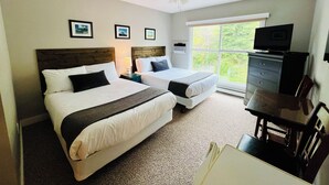 Ein Hotelzimmer mit zwei Betten, einem Deckenventilator, einem Fernseher auf einer Kommode und einem Fenster mit Blick auf Grünflächen.