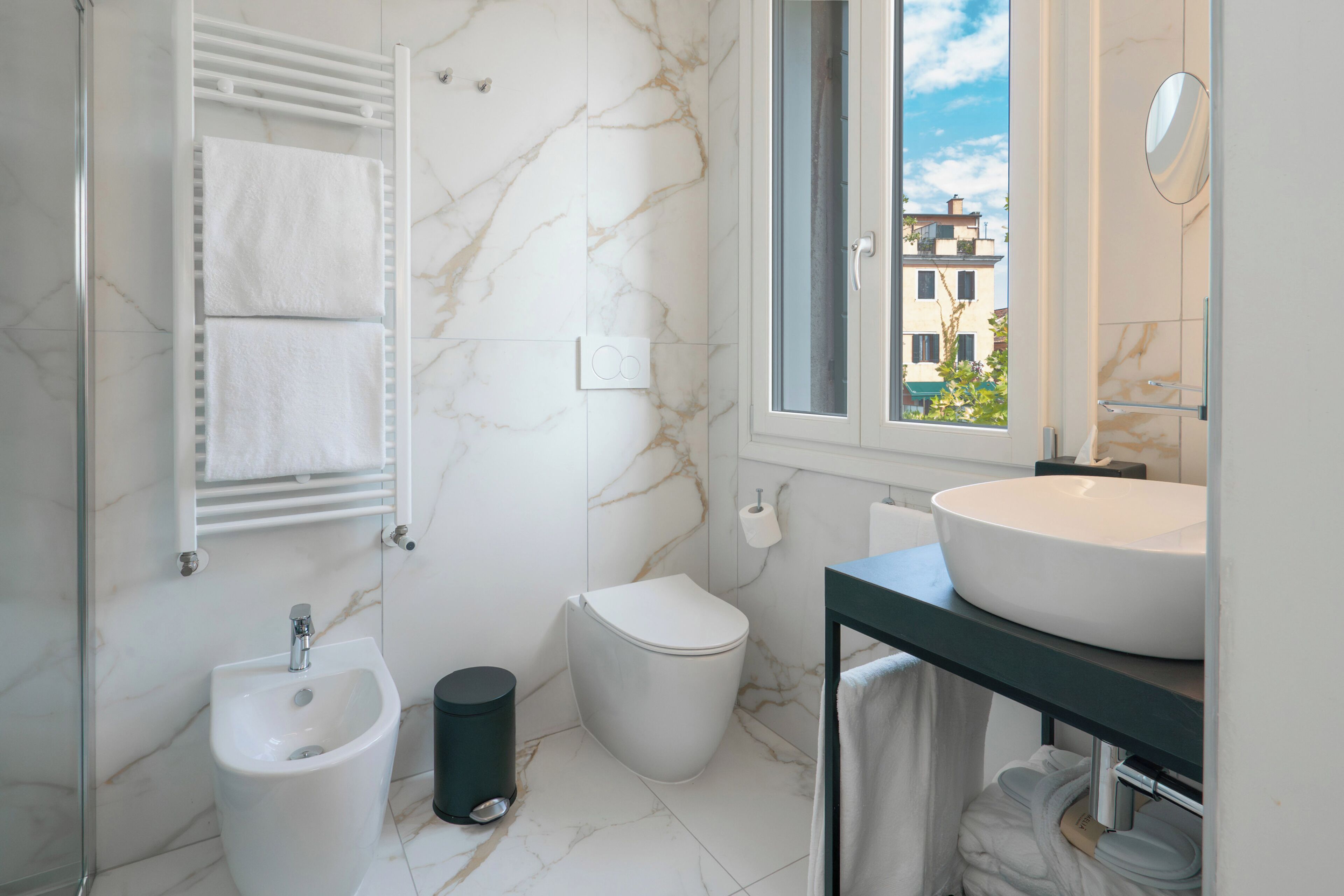 Une salle de bain moderne équipée d’un bidet, de toilettes et d’un lavabo, avec des murs en marbre et une fenêtre donnant sur des immeubles.