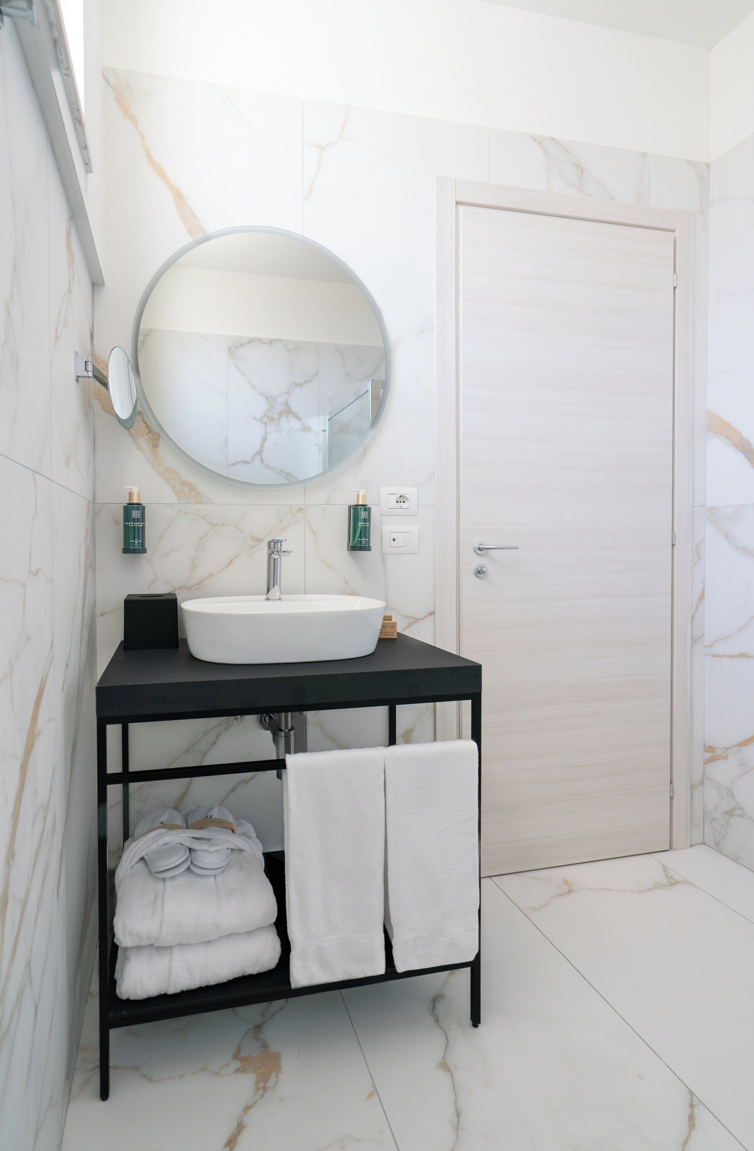 A bathroom with a white marble sink, black vanity, and a round mirror. There are two green bottles on the wall, a towel rack with folded towels, and a white door.