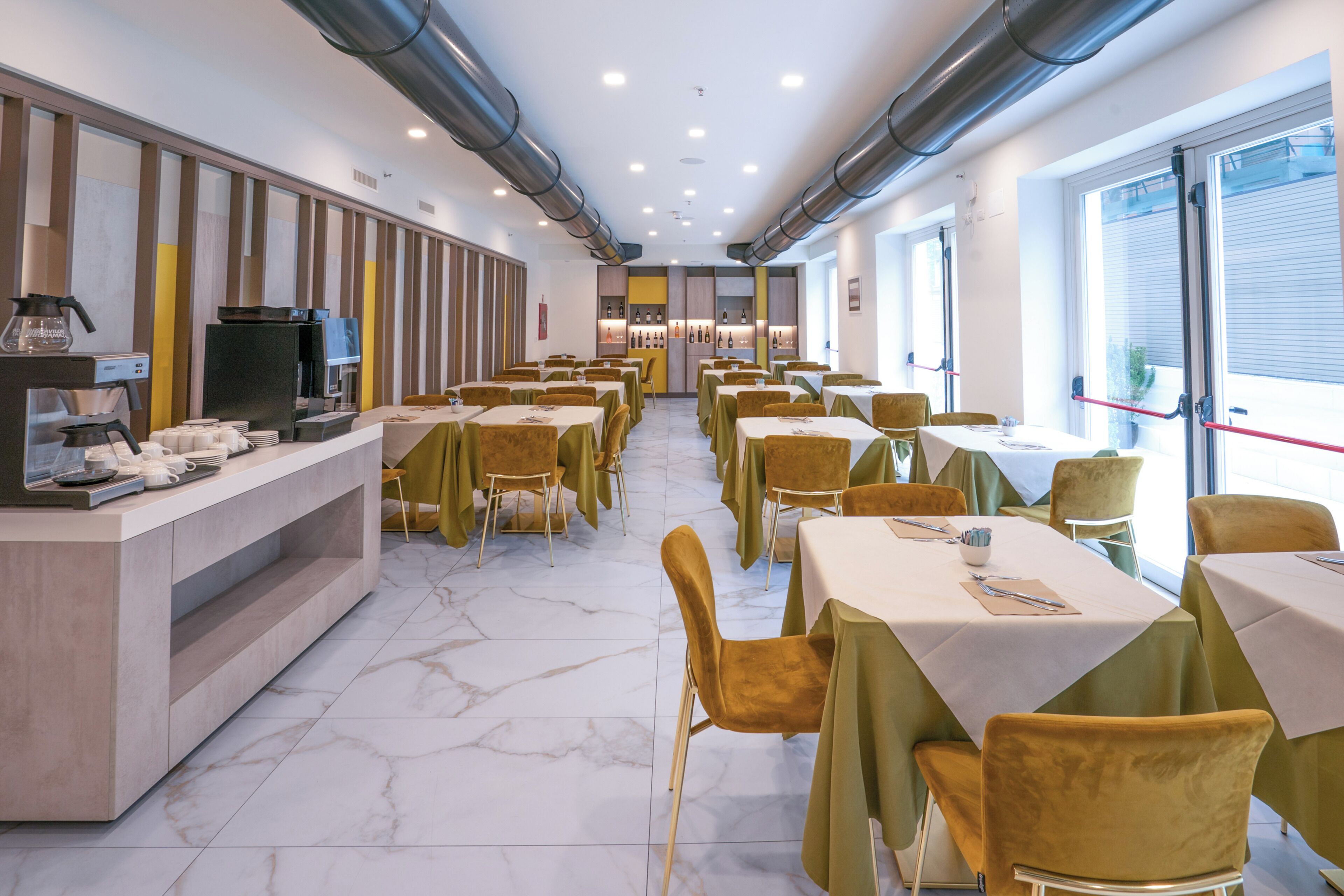 A modern dining area with marble flooring, green-covered tables and chairs, and a central counter with a coffee machine.