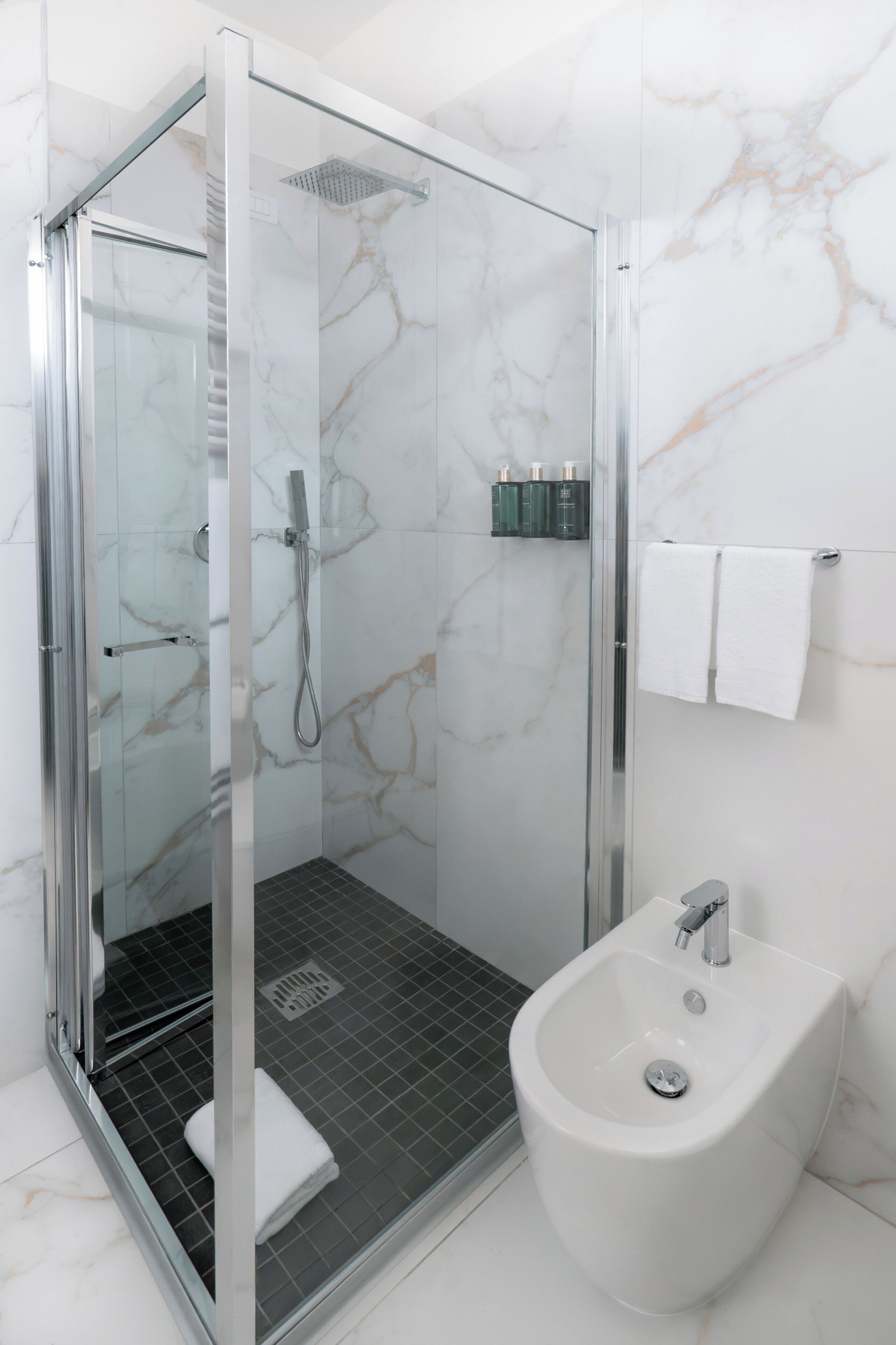 A modern bathroom with a glass-enclosed shower, a white pedestal sink, and marble walls.