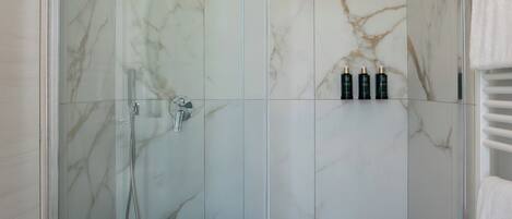 Una ducha sin barreras con puerta de vidrio, paredes de mármol y suelo de baldosas negras. Hay tres botellas en la pared y una toalla en el suelo.