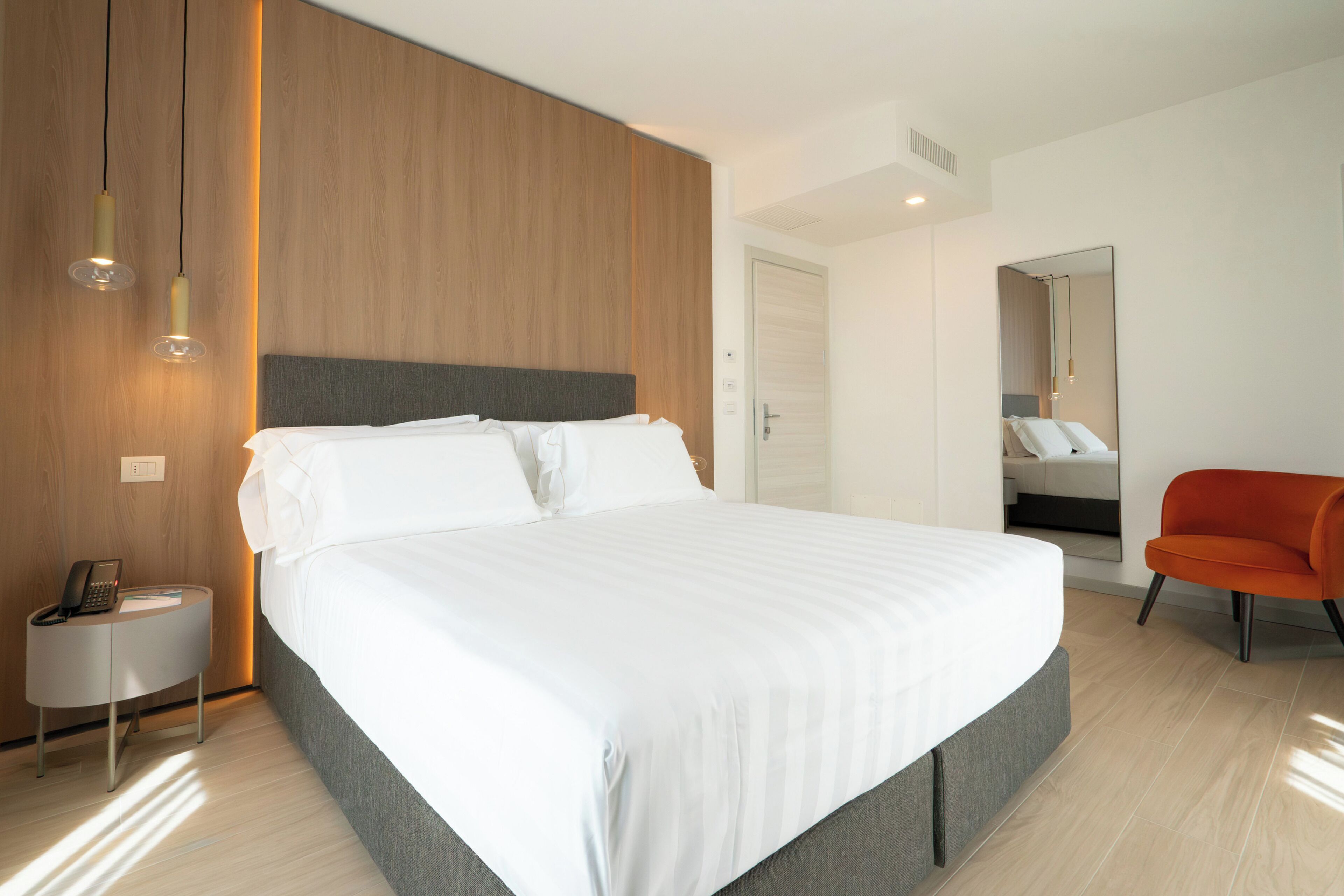 A modern hotel room with a large bed, a bedside table with a phone, and a bright orange chair.