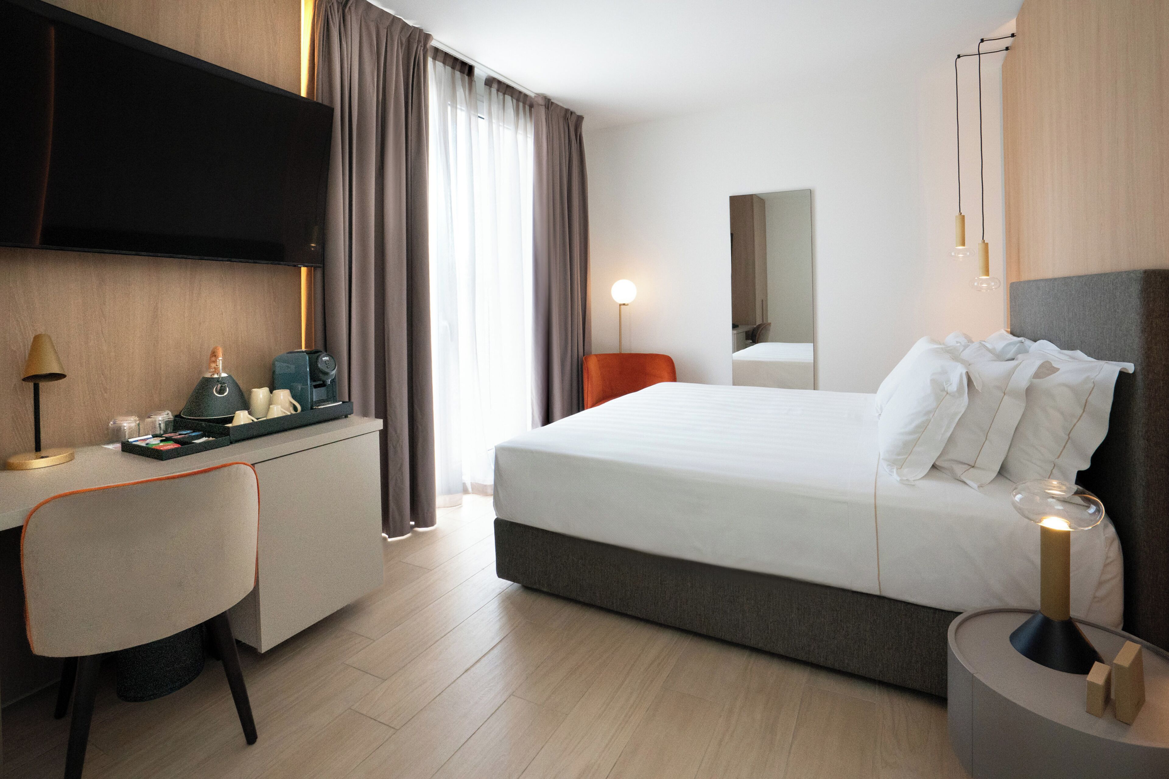A modern hotel room with a bed, a desk with a lamp, a TV, and a bedside table with a lamp.