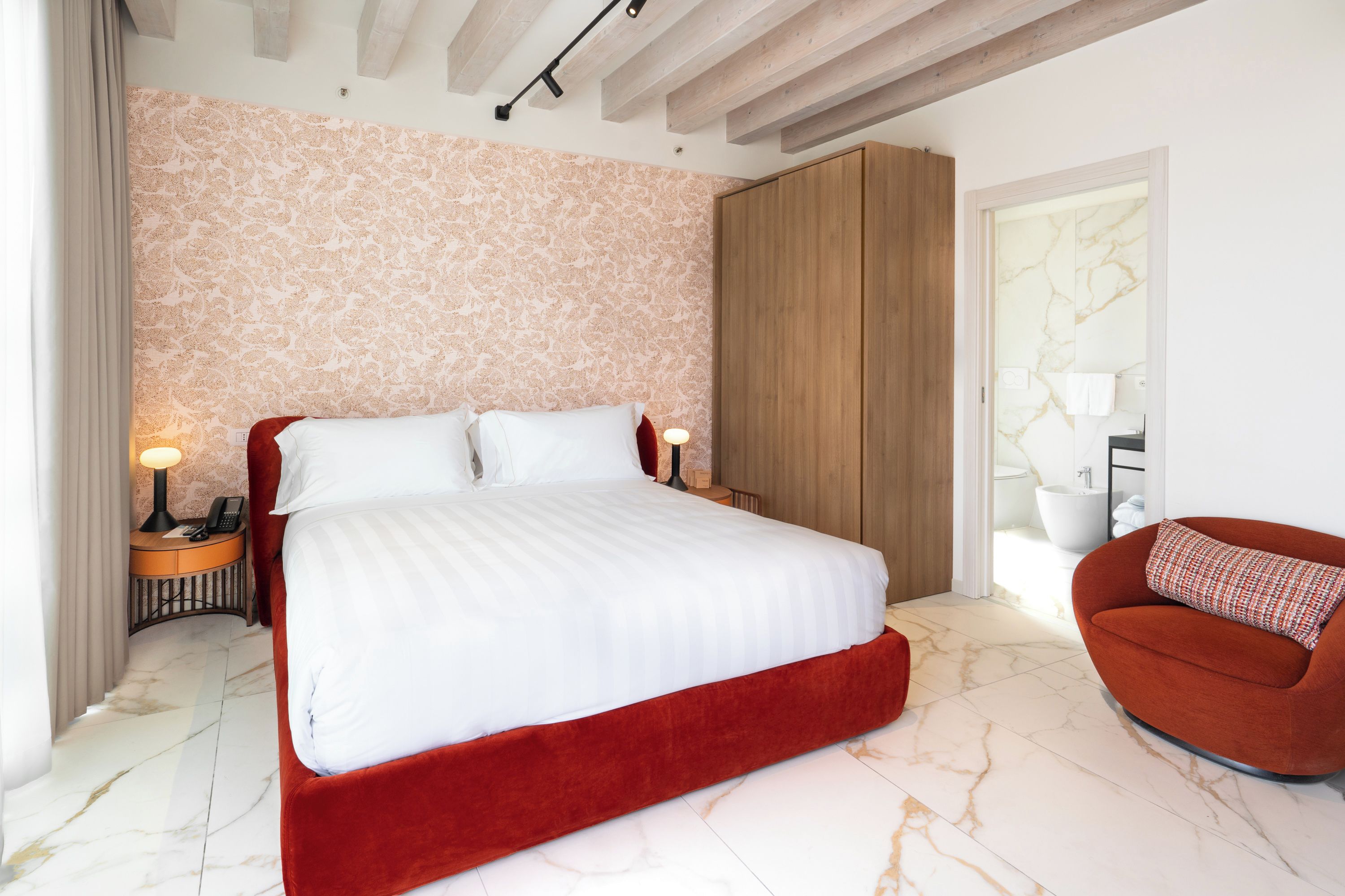 Une chambre d’hôtel avec un grand lit, une armoire en bois, un fauteuil rouge et une salle de bain avec un sol en marbre.