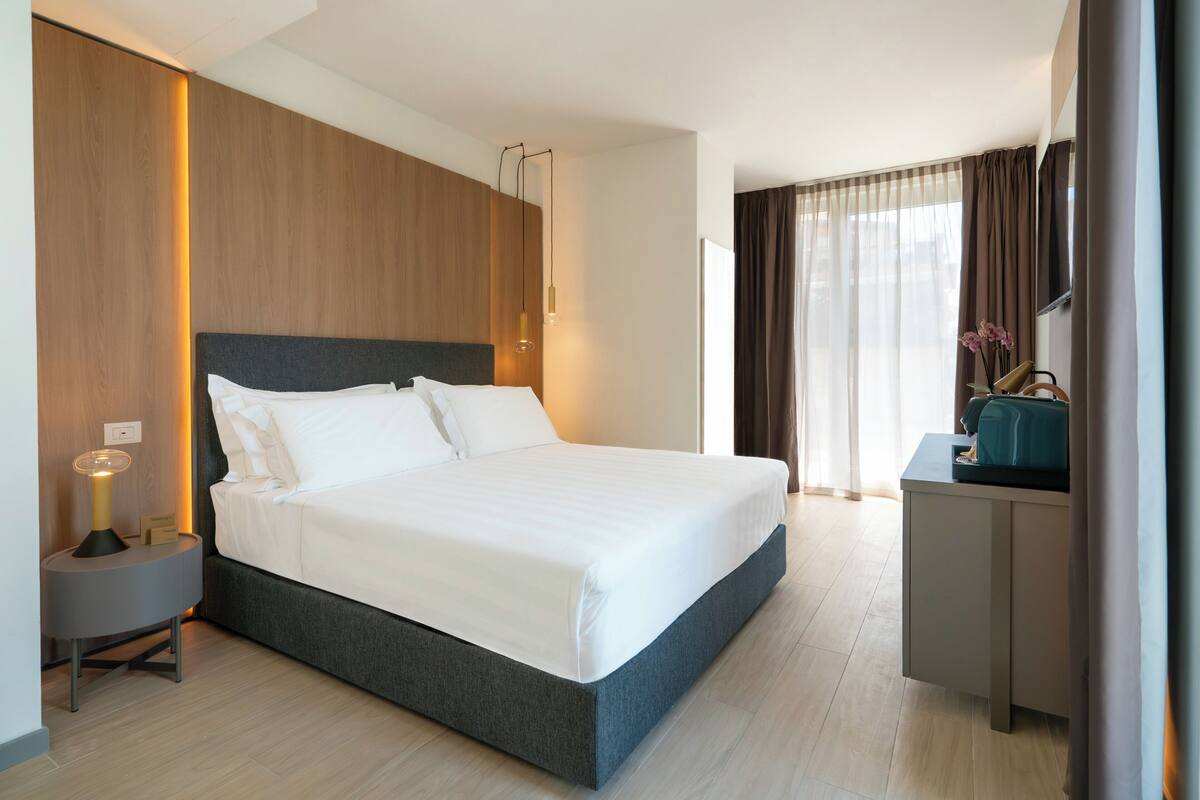 Une chambre d’hôtel moderne dotée d’un grand lit, d’une table de chevet avec une lampe, d’un bureau avec une chaise et offrant une vue sur l’extérieur à travers des rideaux légers.